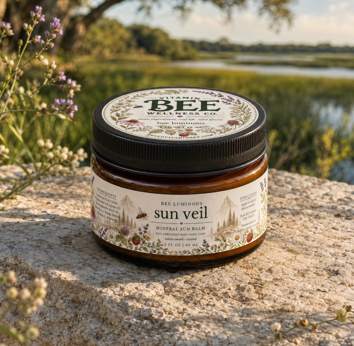 A jar of Vitamin Bee Naturals 'Lime Balmistone' Mineral Sun Balm sits on a stone surface outdoors with a lake and trees in the background. The product has a black screw-top lid and a cream-colored label with botanical illustrations.