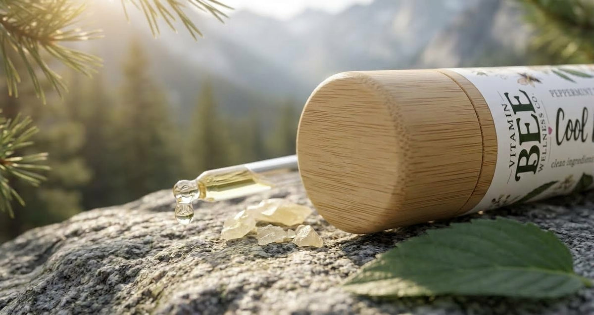 A premium custom bamboo lip balm tube resting horizontally on a rugged granite rock outdoors, highlighting the natural wood grain casing with a clean, unengraved cap. The tube is surrounded by fresh peppermint sprigs, a clear glass dropper with jojoba oil, and shards of raw cocoa butter against a sunlit alpine mountain background.